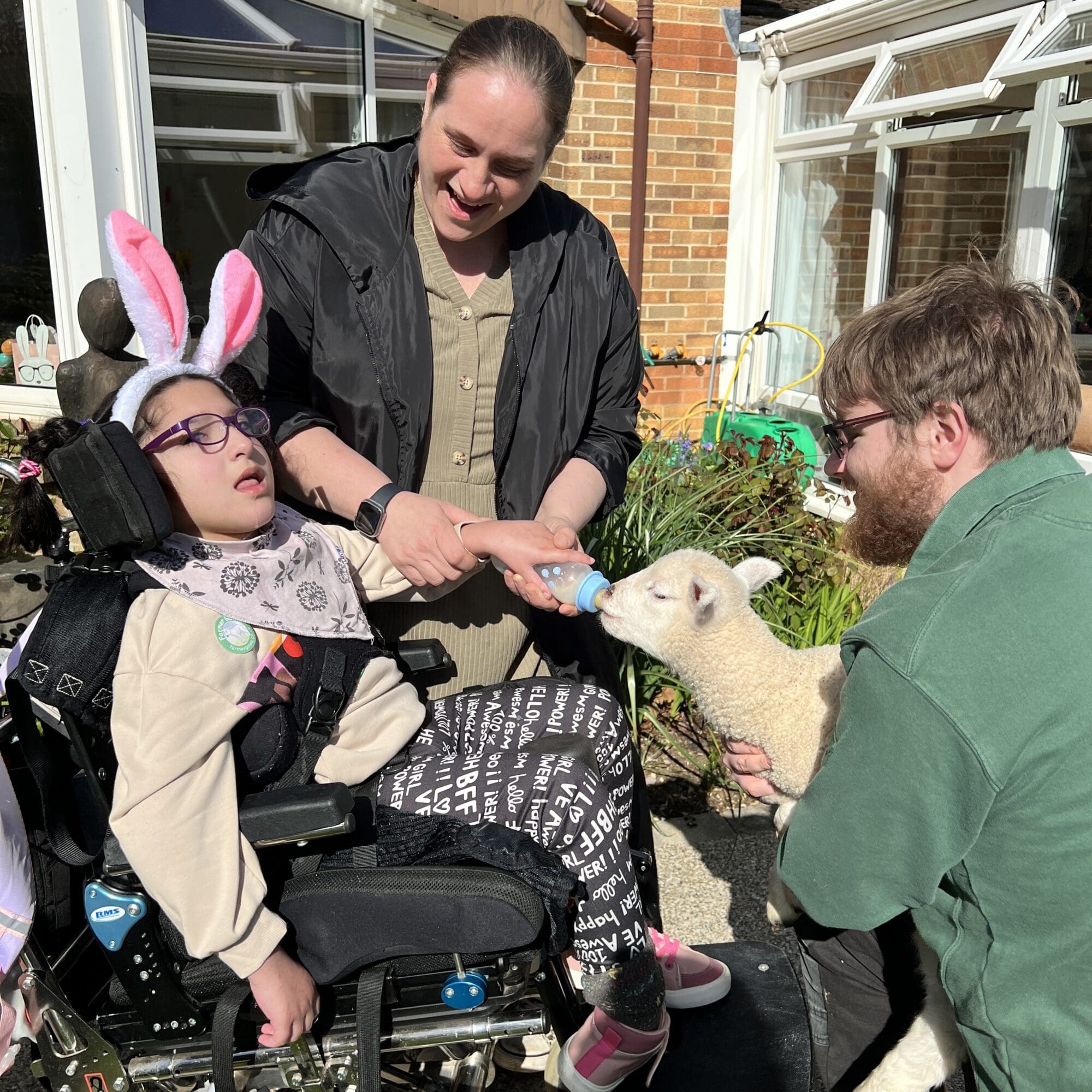a girl in a wheelchair with a woman standing over her feeding a lamb who is being held by a man in a green top